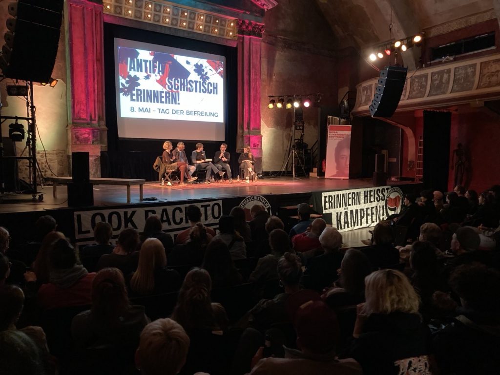 Man sieht ein vollbesetztes UT Connewitz, ein altes Kino, mit einem Podium mit 5 sprechenden Menschen sowie verschiedenen Transparenten, ua.a. Erinnern heißt kämpfen