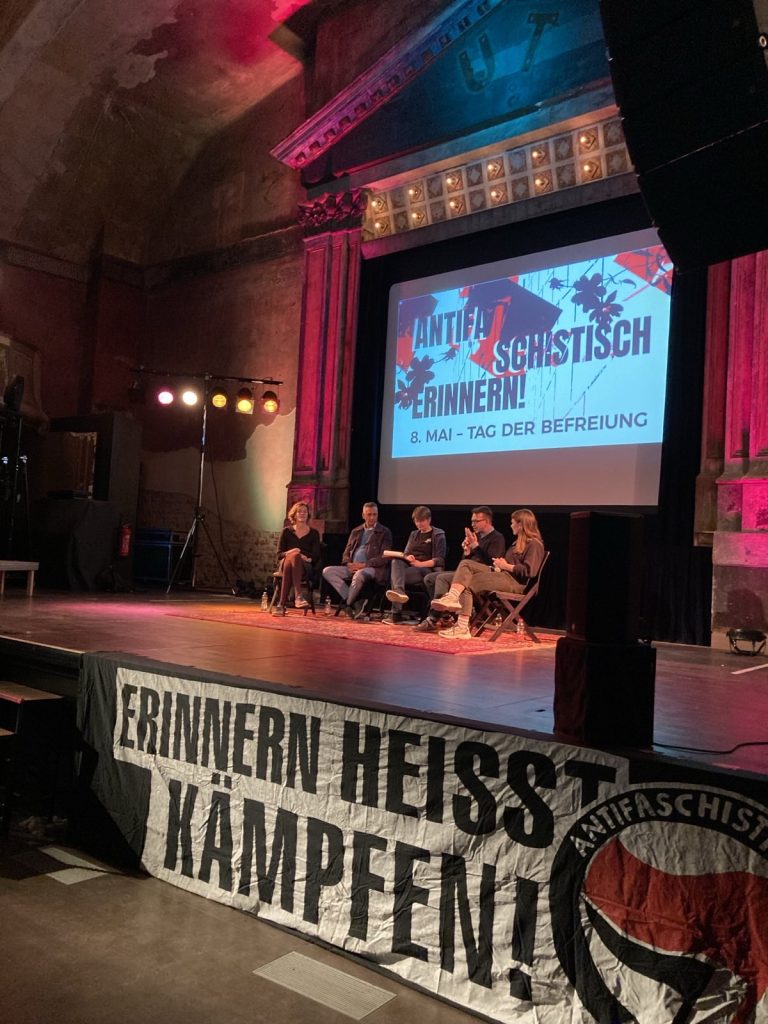 Bühne eines alten Kinos mit fünf Menschen, die unter einem Bild mit Antifaschistisch erinnern - 8. Mai Tag der Befreiung sitzen